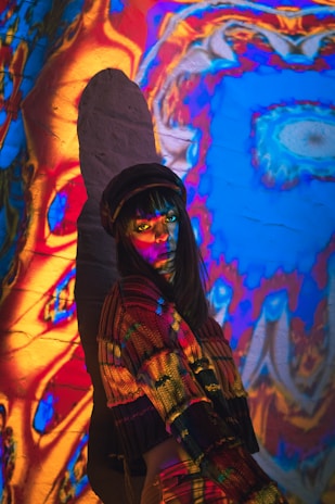 A person stands against a wall covered with psychedelic, colorful projections. The individual is wearing a multicolored sweater and a hat, with vibrant colors such as red, blue, and yellow cast onto them. Shadows and light interact to create an abstract and vivid atmosphere.