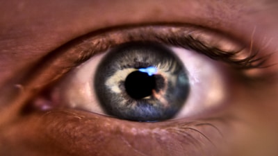 A close-up of a human eye, focusing on the iris and pupil. The iris has a mixture of blue and gray hues with intricate patterns and reflections of light visible. The eyelashes are also partially visible around the eye.