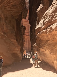 A narrow canyon with towering rock formations leads to a glimpse of an ancient architectural structure. The scene is filled with several people walking and taking photos, suggesting it is a popular tourist site. The sunlight casts dramatic shadows on the rocky walls.