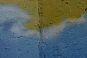 A textured wall with a combination of faint blue and yellow paint. There is a vertical crack running through the middle of the wall. The paint appears to have been applied unevenly, with some areas blending together.