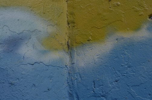 A textured wall with a combination of faint blue and yellow paint. There is a vertical crack running through the middle of the wall. The paint appears to have been applied unevenly, with some areas blending together.