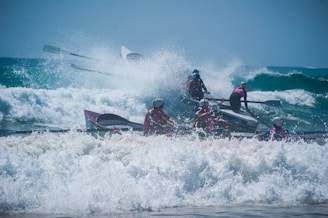 A group of people in helmets are rowing a boat through strong waves in the ocean. The water is crashing around them, creating splashes and foam. They appear to be participating in a coordinated rowing activity or race.