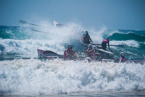 A group of people in helmets are rowing a boat through strong waves in the ocean. The water is crashing around them, creating splashes and foam. They appear to be participating in a coordinated rowing activity or race.