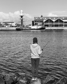 A person is standing on rocks, facing a body of water and holding a fishing rod. In the background, there is a harbor scene with a lighthouse and industrial buildings.