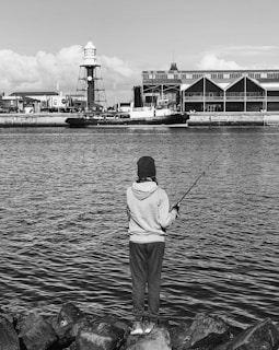 A person is standing on rocks, facing a body of water and holding a fishing rod. In the background, there is a harbor scene with a lighthouse and industrial buildings.