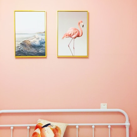 A room with a pale pink wall features two framed pictures; one depicts ocean waves, and the other shows a pink flamingo. Below the pictures, a white metal bed frame is visible with a pillow that also has a flamingo design.