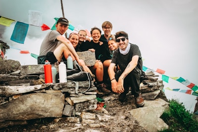 A group of seven people are gathered on a rocky mountain top, smiling and posing for a photo. Colorful prayer flags are strung in the background, suggesting a high-altitude Himalayan setting. Some individuals are holding reusable water bottles, and one person has a sign with 'Yanjing Ri' written on it.