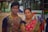 A young man and a middle-aged woman are standing side by side in front of a colorful bus. The young man is wearing glasses and a shirt with a pattern of stars, while the woman is dressed in a traditional sari with a red and gold patterned design. The background shows a blurry outdoor scene with another person and some buildings.