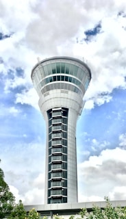 A tall and sleek control tower stands prominently against a background of partly cloudy skies. The modern design features a cylindrical shape with a wide observation deck near the top. The structure is surrounded by minimal greenery at the base.