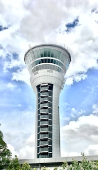 A tall and sleek control tower stands prominently against a background of partly cloudy skies. The modern design features a cylindrical shape with a wide observation deck near the top. The structure is surrounded by minimal greenery at the base.