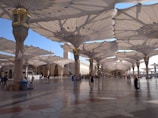 A large open courtyard is shaded by rows of massive, intricately designed umbrella structures. People wearing traditional Middle Eastern attire walk and sit in the area. The architecture includes ornate columns and a distant view of a minaret, creating an atmosphere of elegance and cultural significance.
