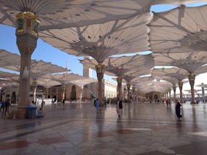 A large open courtyard is shaded by rows of massive, intricately designed umbrella structures. People wearing traditional Middle Eastern attire walk and sit in the area. The architecture includes ornate columns and a distant view of a minaret, creating an atmosphere of elegance and cultural significance.