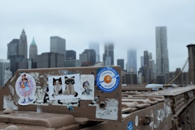 A section of a bridge is adorned with various colorful stickers, while the background features a skyline with tall buildings partially obscured by fog. The stickers include cartoon characters, two cats, and a basketball-themed sticker. The bridge structure has a weathered look with visible bolts and slight graffiti.