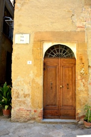 A rustic wooden door set within a stone wall, with an arched window above. The wall appears aged, with faded plaster and patches revealing underlying brickwork. A small street sign reads 'VIA PIA' beside the door, and a house number '1' is displayed. Two potted plants are placed on either side, adding a hint of greenery.