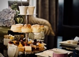 A tiered cake stand displaying a variety of pastries and scones. The top tier contains scones, while the lower tier features desserts garnished with fruits and chocolate. There's a coffee cup set on the table alongside a teapot. In the background, a vase with hydrangeas adds a decorative touch.