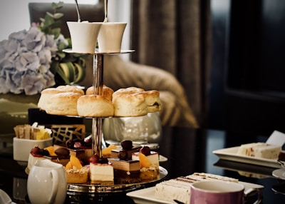 A tiered cake stand displaying a variety of pastries and scones. The top tier contains scones, while the lower tier features desserts garnished with fruits and chocolate. There's a coffee cup set on the table alongside a teapot. In the background, a vase with hydrangeas adds a decorative touch.