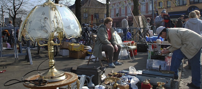 A flea market scene with various items for sale including a decorative brass lamp with a fancy shade in the foreground. People are browsing through items; a woman sits on a chair while another person bends down to look closely at items. The background shows boxes and various goods displayed on tables and the street. There are bikes and buildings with trees lining the area.