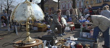 A flea market scene with various items for sale including a decorative brass lamp with a fancy shade in the foreground. People are browsing through items; a woman sits on a chair while another person bends down to look closely at items. The background shows boxes and various goods displayed on tables and the street. There are bikes and buildings with trees lining the area.