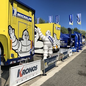 A large yellow truck adorned with the Michelin logo is parked alongside several blue tent structures. The iconic Michelin Man, in white, waves from the side of the truck. Banners and flags bearing the Michelin branding are visible, along with a 'Kronos Racing' sign in the foreground. The scene appears to be set outdoors on a sunny day.