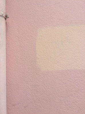 A textured wall painted in a light pink hue with a subtle patch of slightly different color, possibly an attempt at color matching. To the left, there is a vertical cylindrical object that appears to be a pipe, also painted in a similar pink shade.
