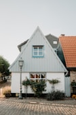 A quaint, small house with an A-frame roof and a light blue siding. The house features two windows, one on the second story with blue trim and one on the ground floor adorned with flowerboxes filled with greenery. A vintage-style street lamp stands in front of the house on a cobblestone pathway, with additional houses visible on either side. The roof of an adjacent house is made up of orange tiles.