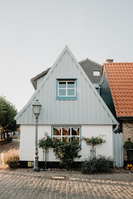 A quaint, small house with an A-frame roof and a light blue siding. The house features two windows, one on the second story with blue trim and one on the ground floor adorned with flowerboxes filled with greenery. A vintage-style street lamp stands in front of the house on a cobblestone pathway, with additional houses visible on either side. The roof of an adjacent house is made up of orange tiles.