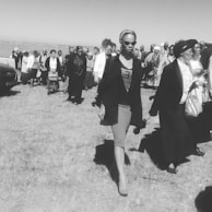 A black and white photograph shows a group of people walking on a grassy field. The central figure is a woman wearing sunglasses, a headband, a dark blazer, and a skirt, who appears to be confident and purposeful. In the background, a diverse crowd moves together, with some women dressed in traditional attire.