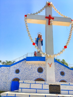 A large, white cross with a rosary made of spherical beads draped around it is featured prominently. At the center of the cross, there is a statue of a woman with a halo, dressed in blue and white, holding a child. The background includes a building with a stone-like pattern painted blue and white and circular windows. There are a few people visible on a platform next to the statue.