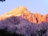 A rugged mountain peak illuminated by the warm hues of sunrise or sunset, casting an orange and pink glow on the rocky surface. The lower portion is shadowed, adding contrast, with patches of snow visible in the crevices. Dark silhouettes of tree branches frame the image.