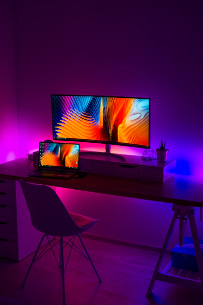 A sleek laptop and desktop setup with colorful peripherals on a modern desk in a bright room.