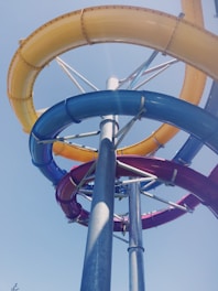 A twisting and vibrant multi-colored water slide structure reaching towards a clear blue sky. The slide features intertwined blue, yellow, and red sections supported by metallic poles, reflecting sunlight.