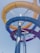 A twisting and vibrant multi-colored water slide structure reaching towards a clear blue sky. The slide features intertwined blue, yellow, and red sections supported by metallic poles, reflecting sunlight.