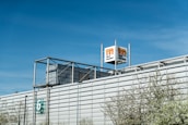 A large industrial building with a modern architectural design, featuring a metallic facade and a prominent logo tower. The building has a number '6' on the side, and some trees with light green spring foliage are visible in the foreground. The sky is clear and blue, indicating a bright day.