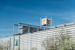 A large industrial building with a modern architectural design, featuring a metallic facade and a prominent logo tower. The building has a number '6' on the side, and some trees with light green spring foliage are visible in the foreground. The sky is clear and blue, indicating a bright day.