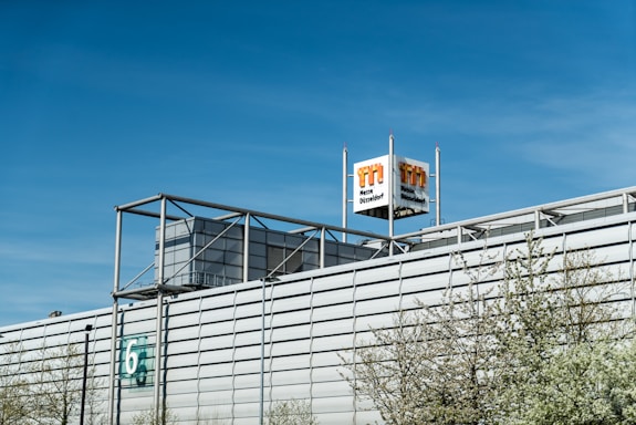 A large industrial building with a modern architectural design, featuring a metallic facade and a prominent logo tower. The building has a number '6' on the side, and some trees with light green spring foliage are visible in the foreground. The sky is clear and blue, indicating a bright day.