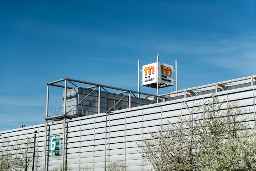A large industrial building with a modern architectural design, featuring a metallic facade and a prominent logo tower. The building has a number '6' on the side, and some trees with light green spring foliage are visible in the foreground. The sky is clear and blue, indicating a bright day.