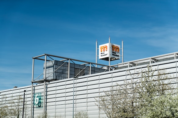 A large industrial building with a modern architectural design, featuring a metallic facade and a prominent logo tower. The building has a number '6' on the side, and some trees with light green spring foliage are visible in the foreground. The sky is clear and blue, indicating a bright day.