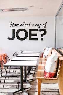 A modern cafe setting with a row of white tables and black chairs arranged neatly. A wooden bench is adorned with decorative cushions featuring colorful designs. Natural light floods in from the right, creating a warm and inviting atmosphere. The wall displays bold black lettering inviting people for coffee.
