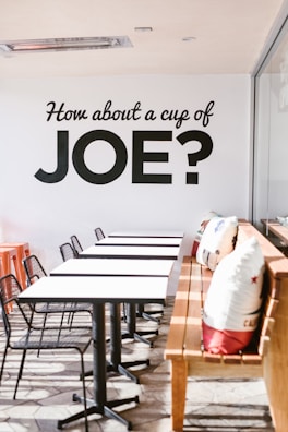 A modern cafe setting with a row of white tables and black chairs arranged neatly. A wooden bench is adorned with decorative cushions featuring colorful designs. Natural light floods in from the right, creating a warm and inviting atmosphere. The wall displays bold black lettering inviting people for coffee.