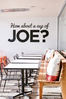 A modern cafe setting with a row of white tables and black chairs arranged neatly. A wooden bench is adorned with decorative cushions featuring colorful designs. Natural light floods in from the right, creating a warm and inviting atmosphere. The wall displays bold black lettering inviting people for coffee.