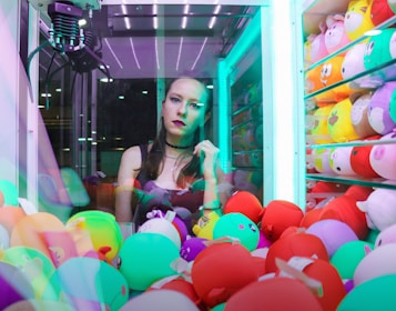 A woman is standing inside a brightly lit claw machine surrounded by colorful plush toys. She is looking directly at the camera with a thoughtful expression. The claw is suspended above the pile of toys, and the background is illuminated with neon lights.