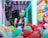 A woman is standing inside a brightly lit claw machine surrounded by colorful plush toys. She is looking directly at the camera with a thoughtful expression. The claw is suspended above the pile of toys, and the background is illuminated with neon lights.