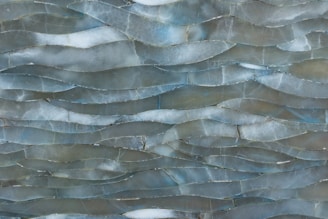 A textured surface composed of overlapping, translucent, grayish-blue tiles with irregular shapes and varied edges. The tiles create a wavy, layered pattern resembling the natural flow of water with subtle variations in color and opacity.