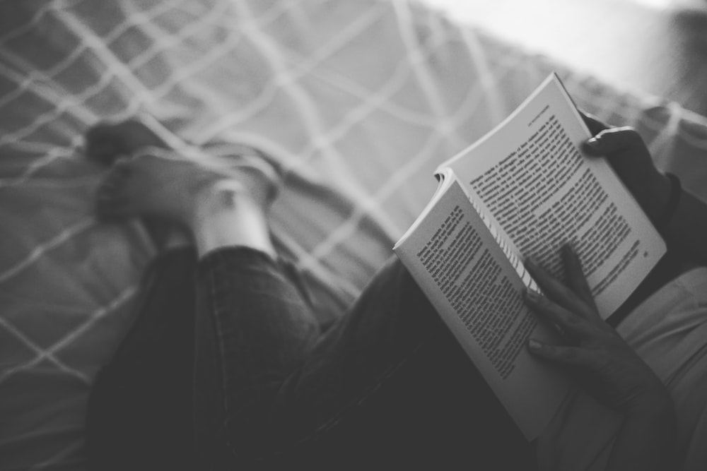 grayscale photography person reading book on top of bed