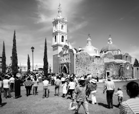 A black and white photo capturing a crowd gathered around a historical stone church with a tall bell tower. The facade is ornately decorated, and several domes are visible. People of various ages are walking and interacting, many wearing hats suggesting a sunny day. Tall, narrow trees and a lamppost are part of the setting.