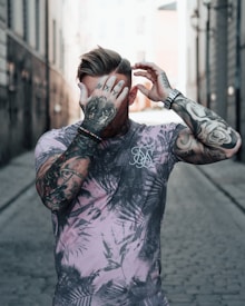 A tattooed person stands in the middle of a narrow cobblestone alley, wearing a patterned t-shirt. They are covering their face partially with one hand, while the other hand adjusts their hair. The background consists of tall buildings with a contrasting bright light at the end of the alley.