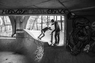 Two people are skateboarding in an urban skate park with graffiti on the walls. The park is under a bridge, casting shadows and creating a moody atmosphere. One person is standing on a ramp holding a skateboard, while the other is performing a trick. Leafless trees and a fence are visible in the background.