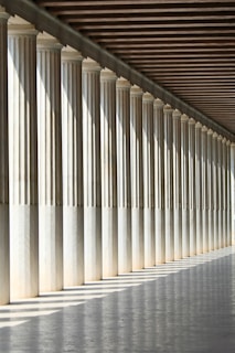 A long row of classical columns creates a sense of symmetry and perspective. The sun casts shadows on the polished floor, adding depth and contrast to the scene. The wooden ceiling complements the classical aesthetic.