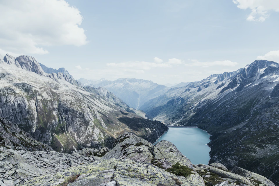 The image depicts a breathtaking view of a mountain range with snow-capped peaks and a deep blue lake nestled within it. The mountains, painted in shades of gray and white, stretch across the horizon, their jagged edges contrasting sharply against the clear sky above. The lake, reflecting the surrounding landscape, is a vibrant shade of blue that mirrors the sky's color. The image is taken from an elevated perspective, offering a panoramic view of the mountains and the lake below.