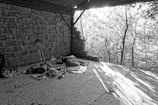 A black and white image depicting an area with improvised camping setup under a roofed structure, against a backdrop of partially visible trees. Camping gear such as a backpack, sleeping bag, trekking poles, and various items are spread on a flat concrete surface. The sunlight filters through the trees, casting shadows on the ground.